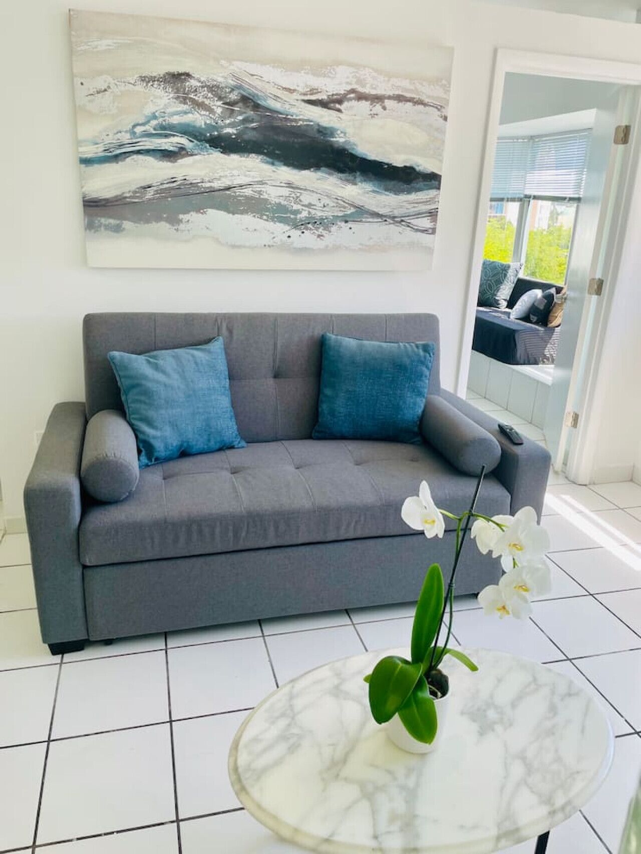 A grey sofa with blue cushions, a white orchid on a marble table, and a large abstract painting on the wall.