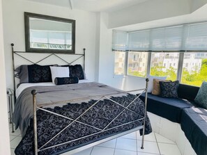 Un dormitorio con cama, ventana con persianas y vistas a edificios, además de una estructura de cama metálica.