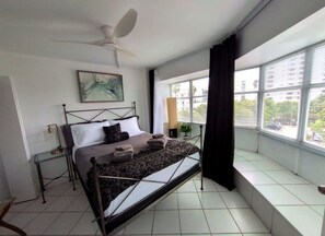 A bedroom with a bed, a ceiling fan, a window with blinds, and a view of buildings outside.