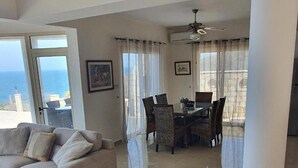 A spacious living area with a sofa, dining table, and chairs, large windows with curtains, and a ceiling fan. - Villa Aquavitus in Pachyammos (Pomos)