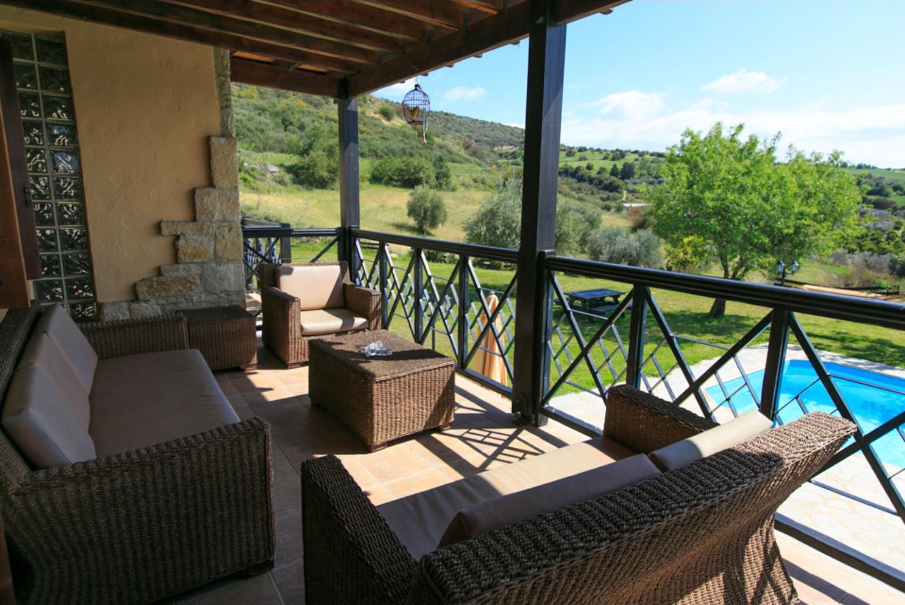 A covered outdoor seating area with wicker furniture, a stone wall, and a view of a swimming pool and hills.