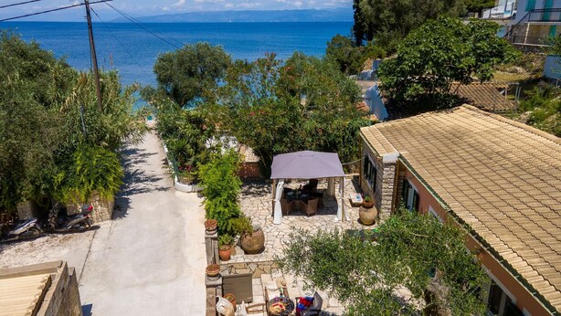Une propriété située en bord de mer, avec un chemin pavé menant à un espace extérieur aménagé, abrité et doté d’une table et de chaises, entouré de plantes en pot et d’un bâtiment en pierre avec un toit en tuiles.