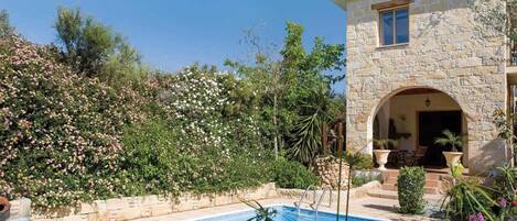 Un edificio de piedra con piscina, rodeado de vegetación exuberante y plantas florecientes.