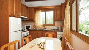 Una cocina con armarios de madera, un refrigerador blanco, un microondas, una estufa y un fregadero. Hay una mesa de comedor con sillas y una ventana con cortinas.