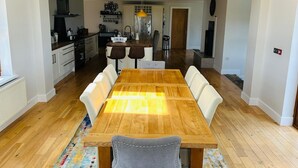 Un vaste espace de salle à manger avec une table en bois et des chaises à revêtement blanc, qui donne sur une cuisine équipée d’appareils modernes et agrémentée d’un luminaire suspendu.