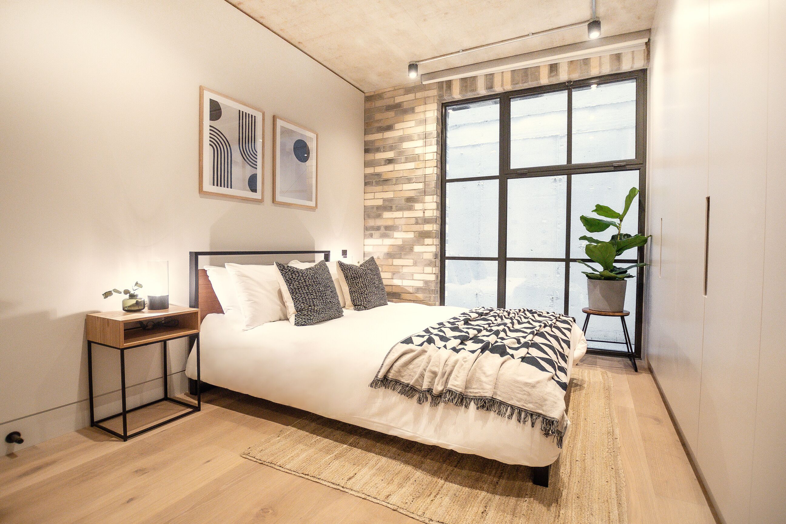 A bedroom with a large bed, a nightstand, a potted plant, and a window with a view.