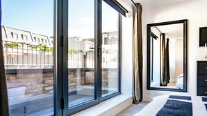 A modern bedroom with large windows, a black and white striped rug, and a mirror on the wall. - Be London - The Bloomsbury Residences (London)