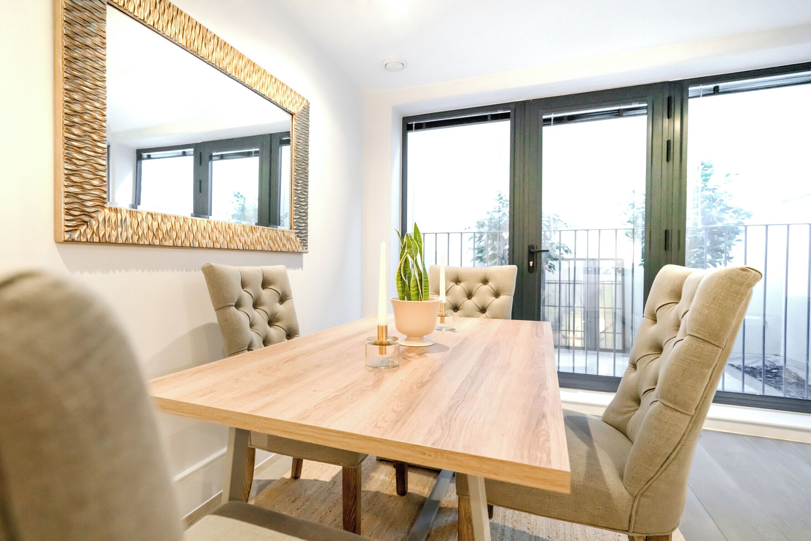 Un comedor con una mesa de madera, sillas acolchadas, un espejo grande y una planta en maceta.
