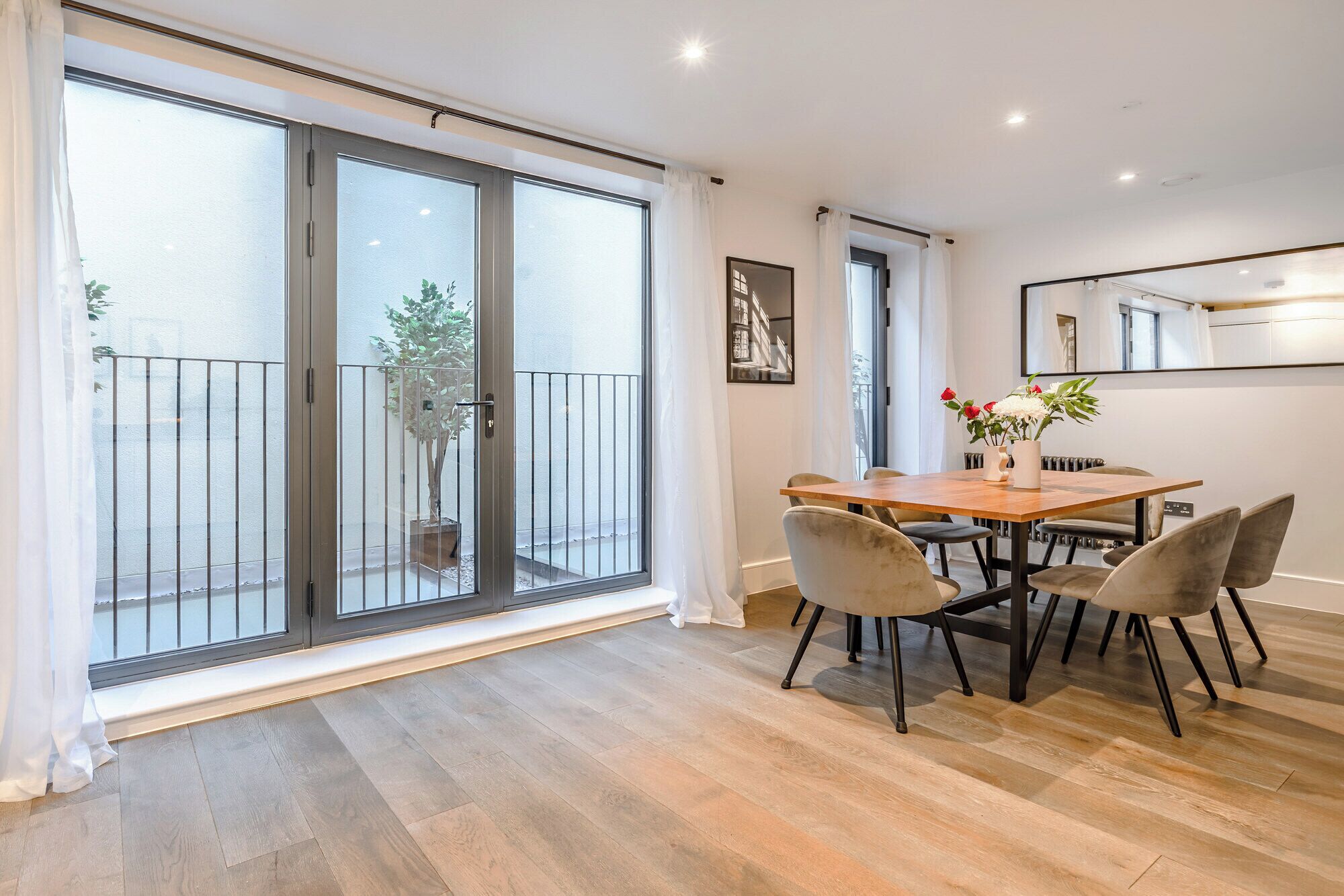 A modern apartment with a wooden dining table, four chairs, a potted plant, and a sliding glass door leading to a balcony.
