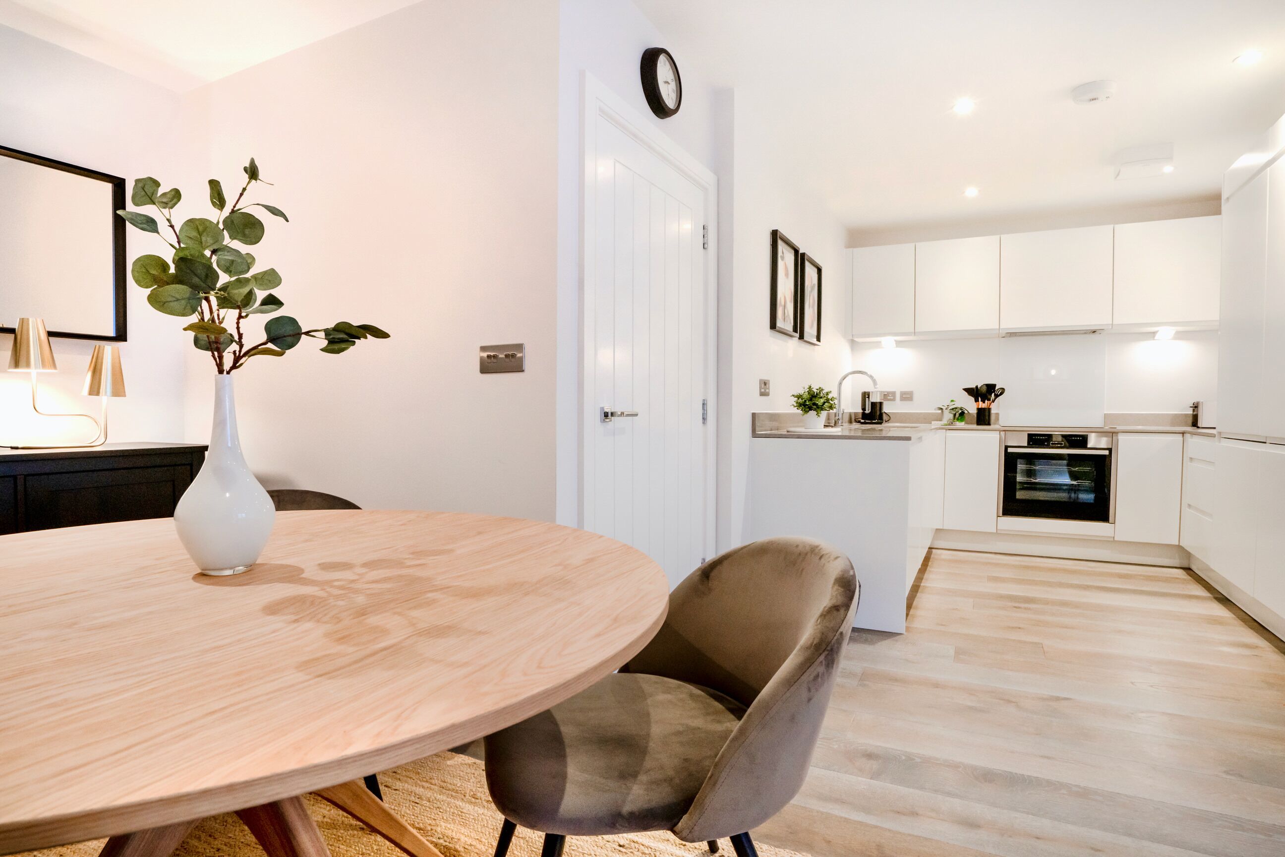 A modern kitchen with white cabinets, a built-in oven, and a dining area with a round wooden table and a single chair.
