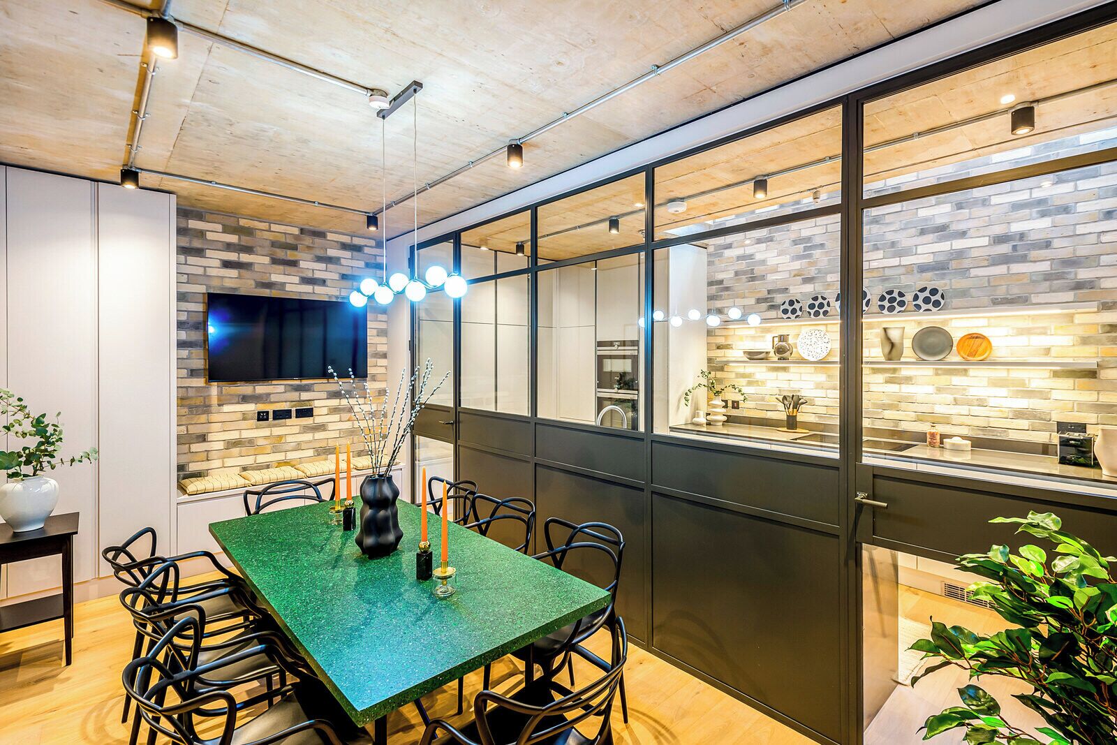 A modern dining area with a green table, black chairs, and a brick wall with a TV.
