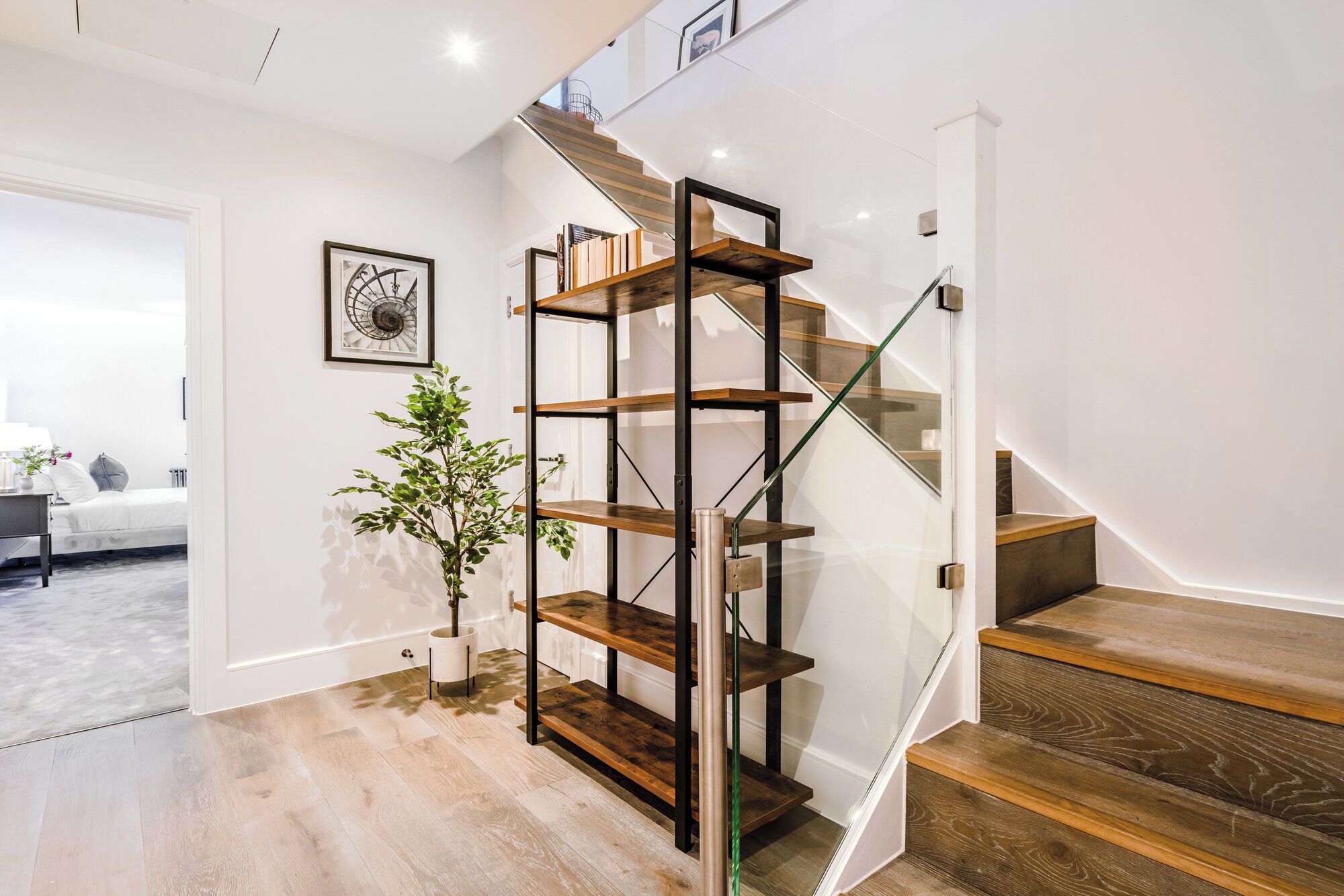A staircase with wooden steps and a glass railing, leading to a landing with a metal shelving unit and a potted plant.
