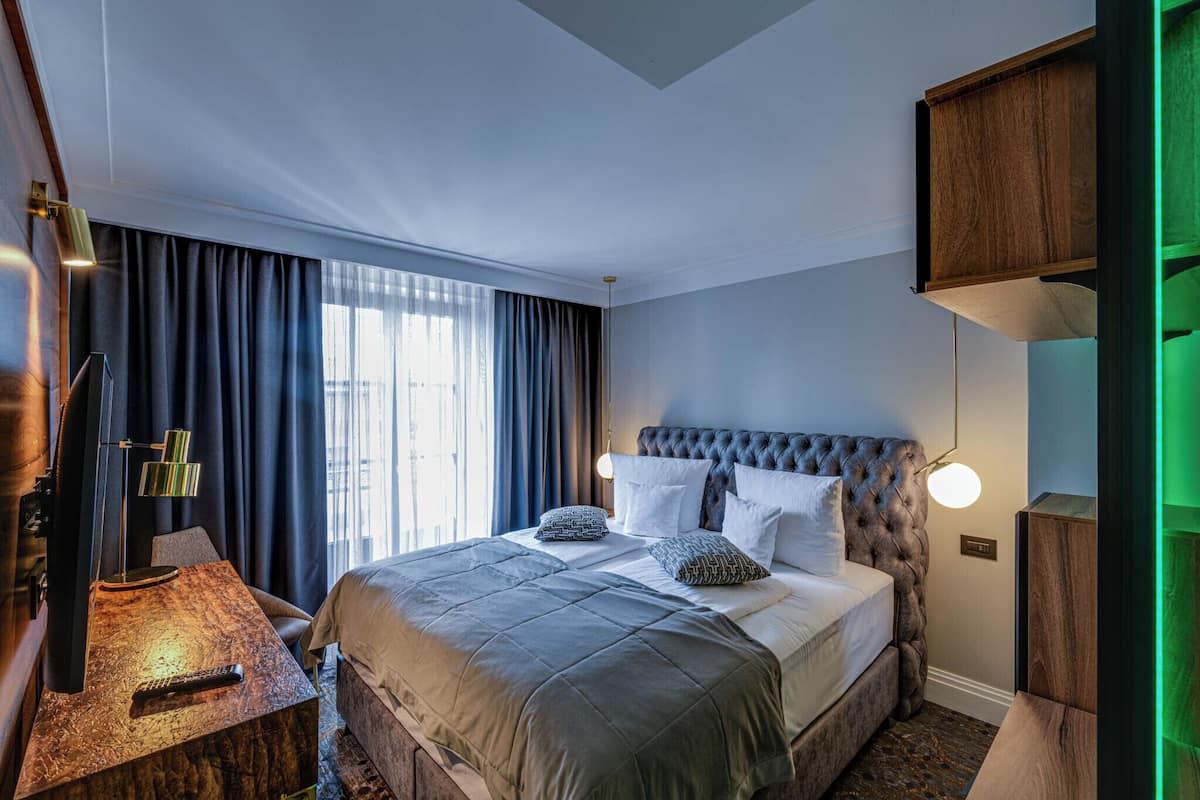 Una habitación de hotel moderna con una cama grande, un escritorio con lámpara y vistas a la ciudad desde la ventana.