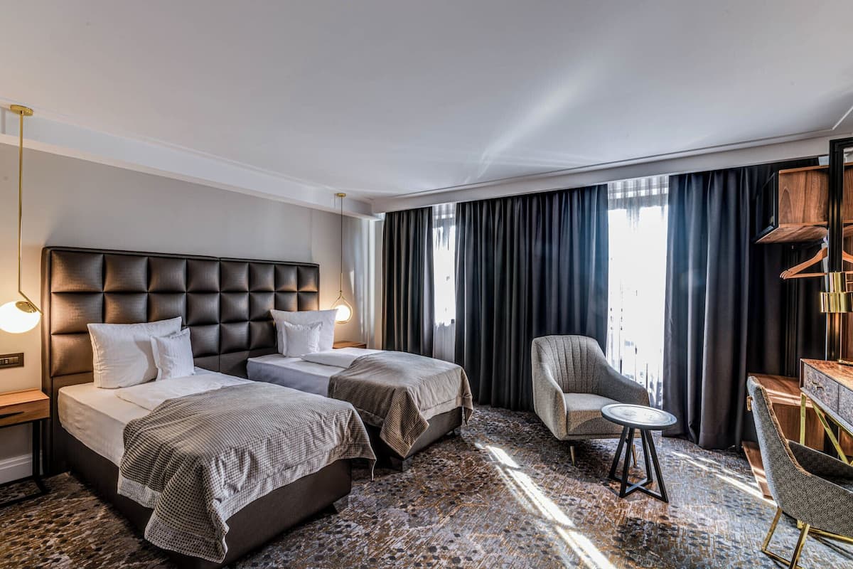 Habitación de hotel con dos camas, un escritorio, una silla y un ventanal con cortinas.