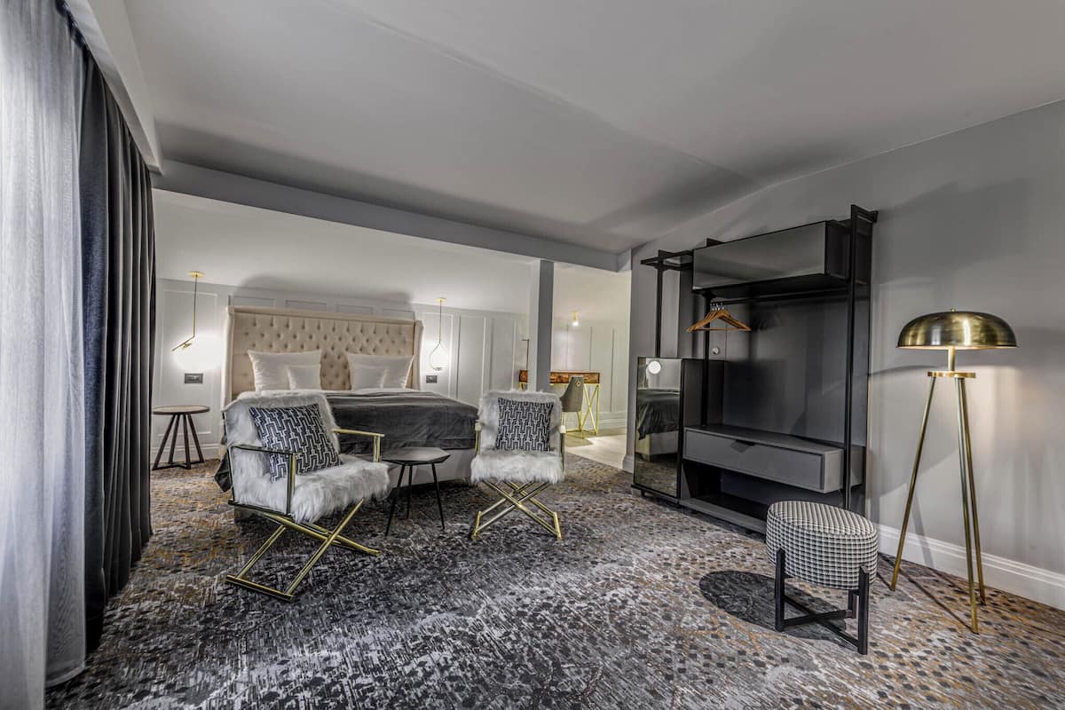 Una habitación de hotel moderna con una cama grande, una zona de estar con sofá y sillones, un televisor de pantalla plana, una mesita auxiliar y una lámpara de pie.