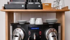 Eine kleine Kaffeestation mit zwei Espressomaschinen, einer Kaffeemühle und einem Regal mit Tassen und einer Kiste.