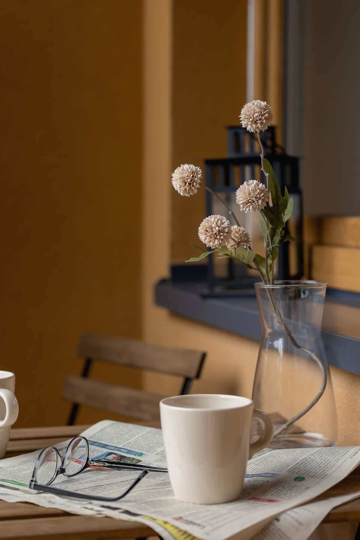 A table with a clear glass vase containing flowers, a white mug, and a pair of glasses on a newspaper.