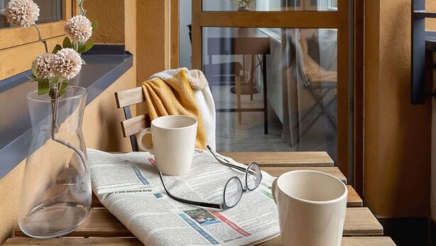 A wooden table with a newspaper, a clear glass vase with flowers, a pair of glasses, and a mug. - Stylish Apartment with Balcony | 2 Bedrooms | Parking. (Kraków)