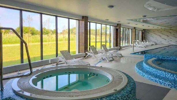 Un espace piscine intérieur moderne, doté d’un jacuzzi, de chaises longues et de grandes fenêtres offrant une vue sur l’extérieur.