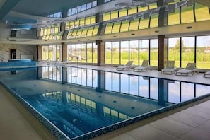 An indoor swimming pool with clear glass walls, lounge chairs, and a view of the outdoors.
