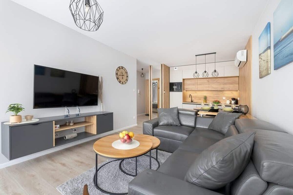 Ekskluzywny salon z aneksem kuchennym. Pomieszczenie jest wyposażone w sofą narożną, TV i wyjście na balkon.