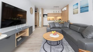 Un salon moderne avec un canapé gris, une table basse en bois sur laquelle on voit des fruits, un téléviseur et, au fond, une cuisine.
