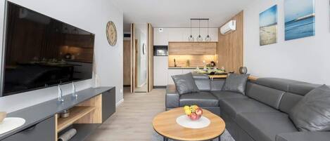 A modern living room with a grey sofa, a wooden coffee table with fruit, a TV, and a kitchen in the background.