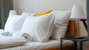 Una cama bien hecha con ropa blanca y una manta amarilla, una lámpara de noche y un cabecero de madera.