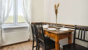 Un comedor con una mesa de madera dispuesta para cuatro personas, flanqueada por sillas negras, un jarrón blanco con plantas secas y una ventana con cortinas blancas.