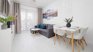 A modern living room with a grey sofa, white dining table, and a small round coffee table. There are potted plants and a painting on the wall.