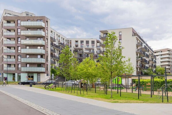 A multi-story residential building with balconies, a paved walkway, and a playground area with trees.