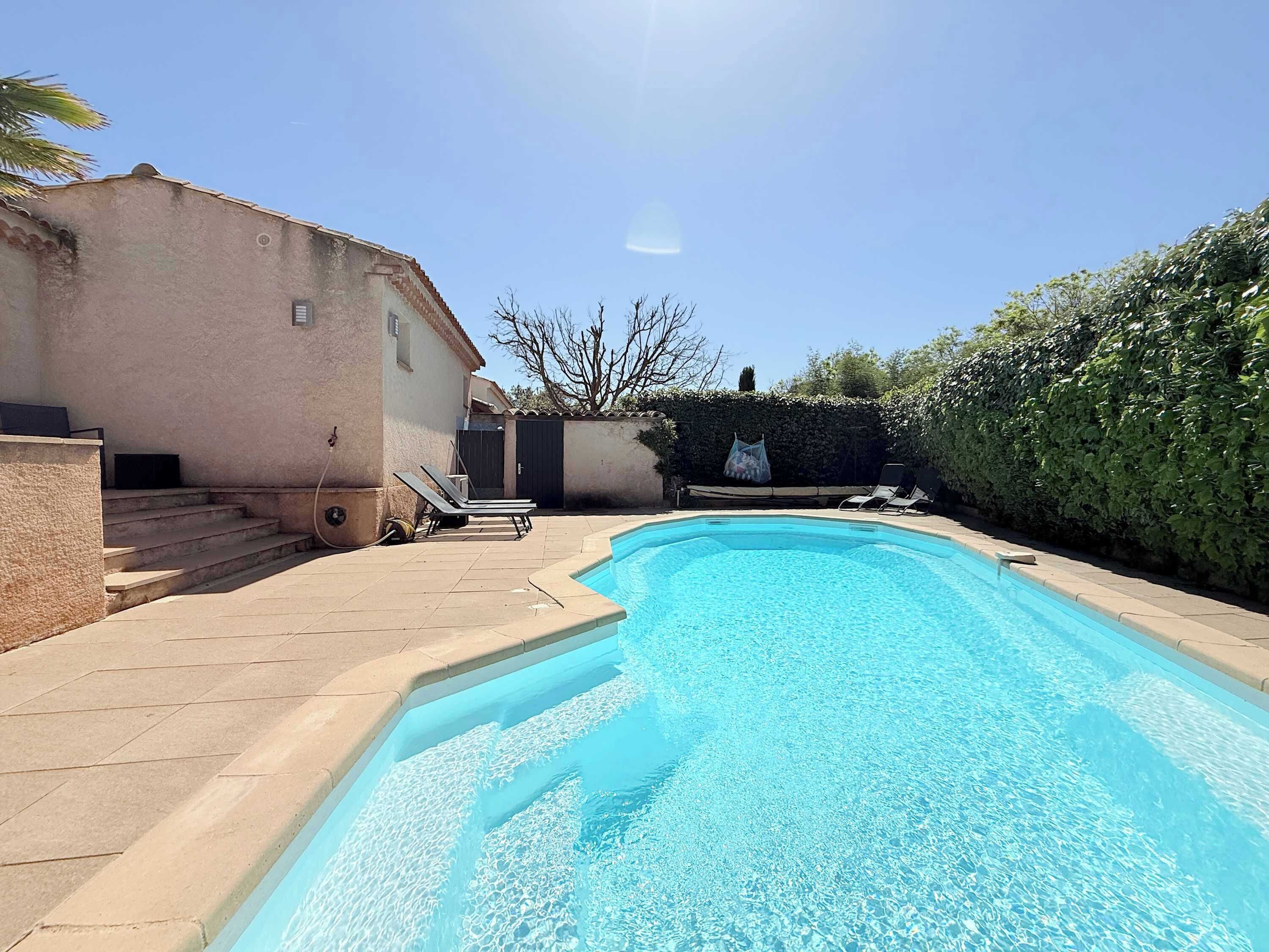 A swimming pool with a tiled deck, a small building, and a hedge.