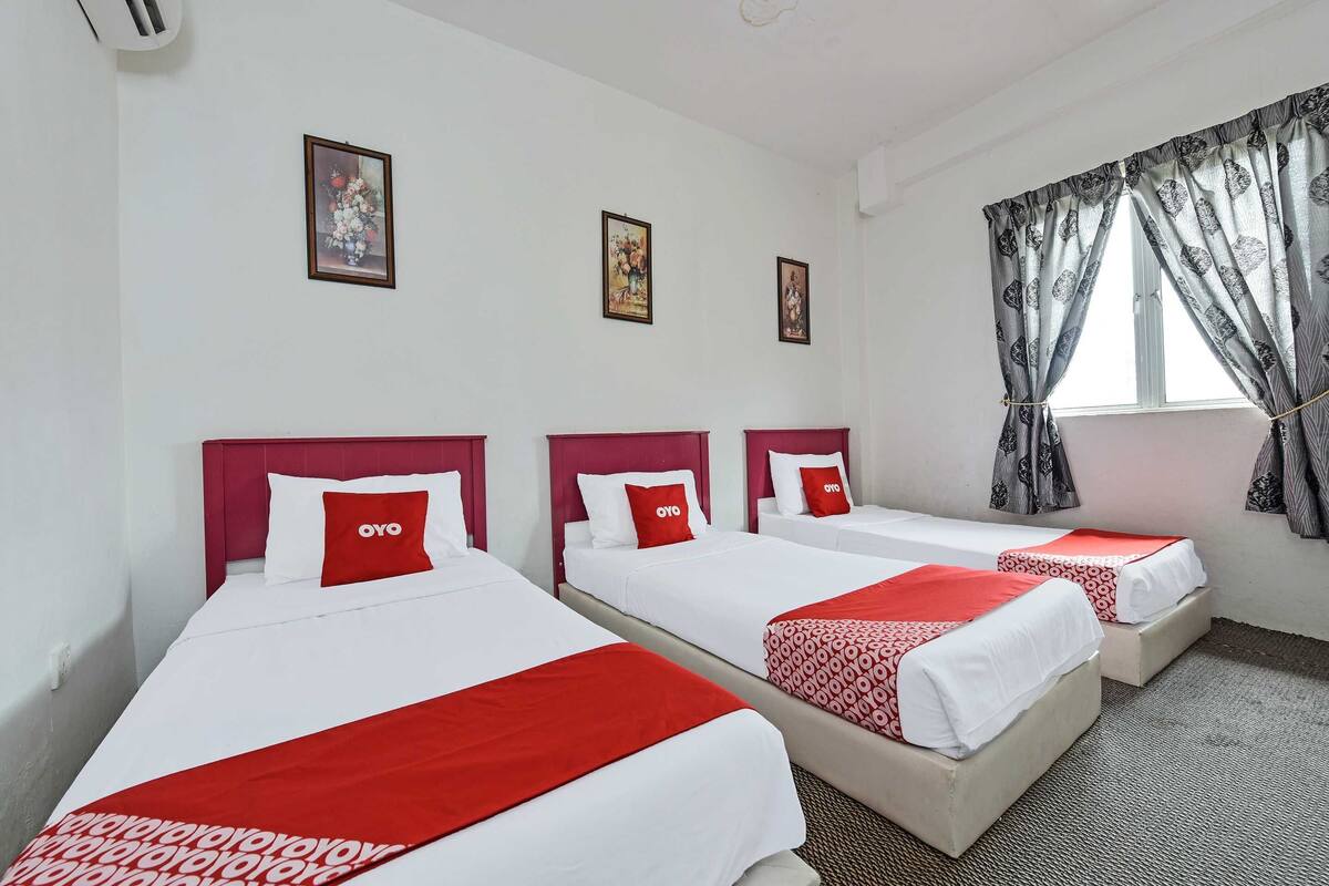 A hotel room with three single beds, each with a red and white bedspread and matching pillows. There are two framed pictures on the wall, a window with curtains, and a carpeted floor.