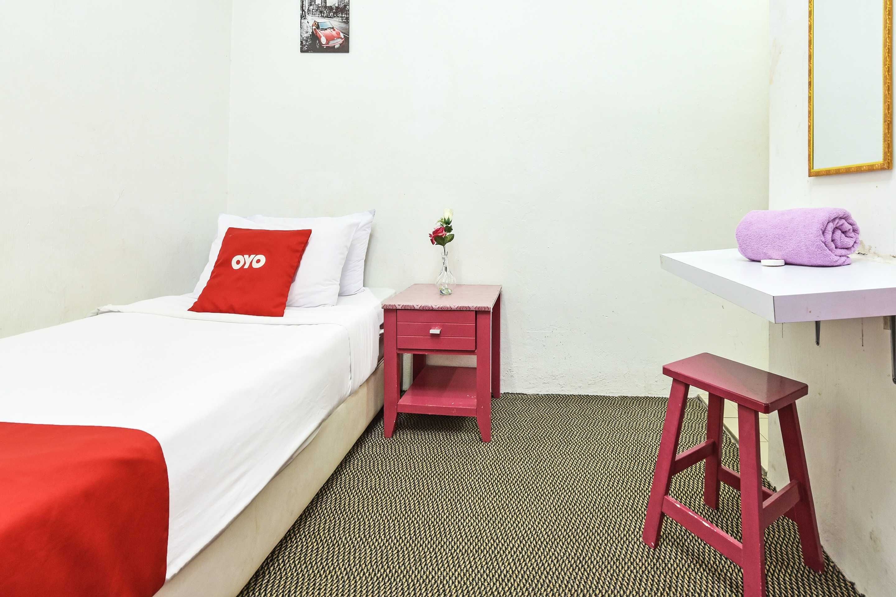 A hotel room with a bed, a small red nightstand with a vase of flowers, a red stool, and a white shelf with a rolled towel.
