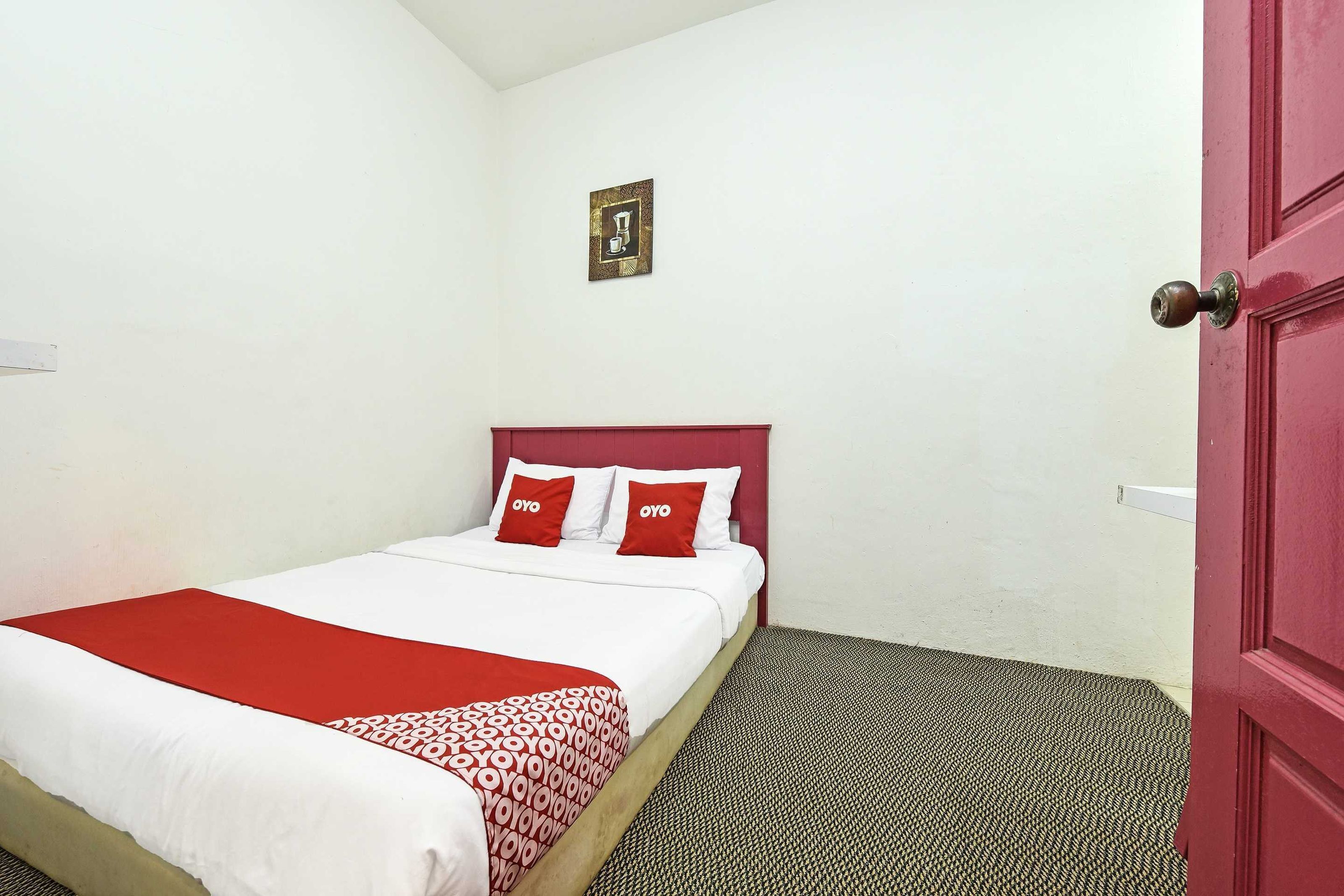 A single bed with a red and white bedspread, two red pillows, and a small framed picture on the wall.