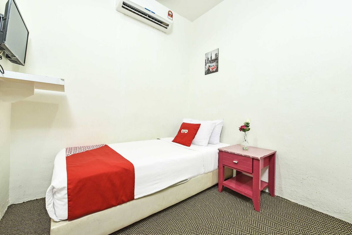 A single-bed room with a red and white bedspread, a small bedside table with a vase of flowers, and a wall-mounted air conditioner.