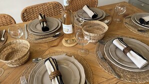 A wooden dining table set with plates, glasses, and a bottle of wine, surrounded by woven baskets and wicker chairs. - La Maison de la Bergère (Fox-Amphoux)