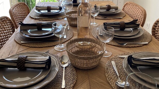 A dining table set with plates, cutlery, glasses, and a bottle, surrounded by wicker chairs and a view of greenery. - La Maison de la Bergère (Fox-Amphoux)