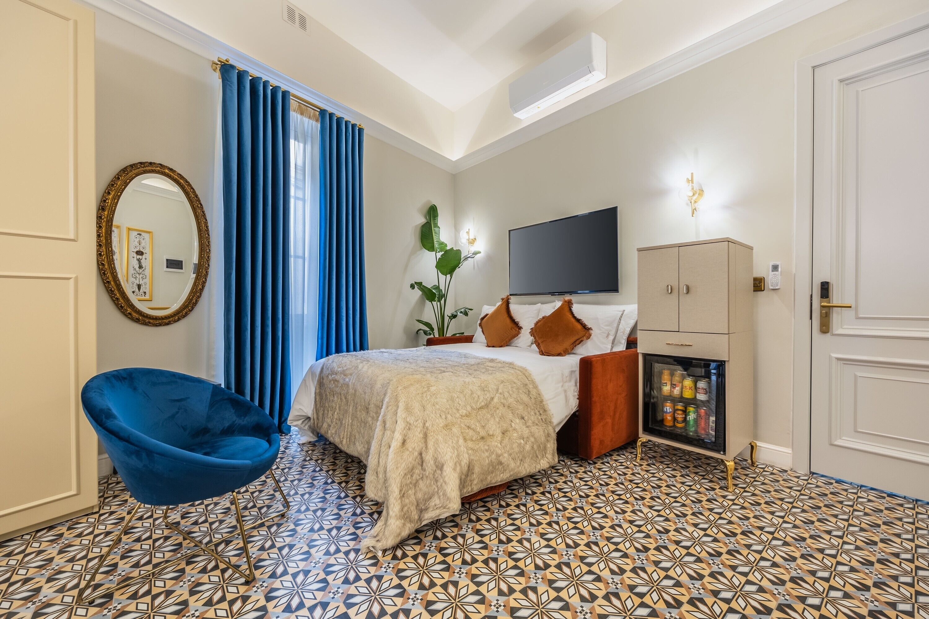 A hotel room with a bed, a blue chair, a television, a minibar, and patterned flooring.