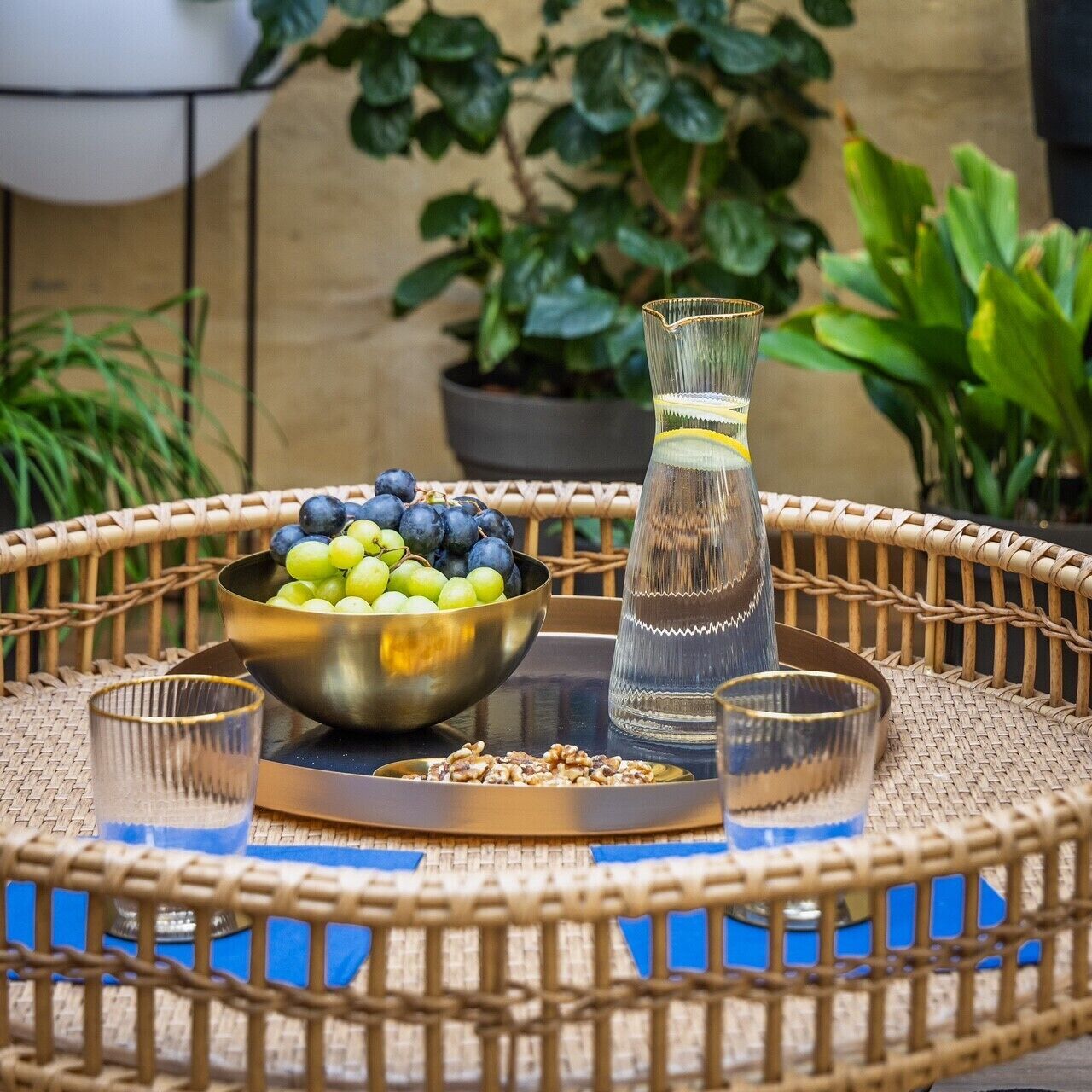 A wicker table with a bowl of fruit, a glass pitcher, and two glasses.