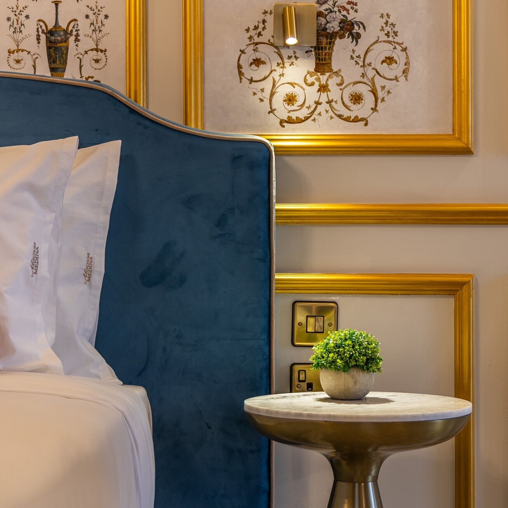 A hotel room with a blue headboard, a bedside table with a potted plant, and a wall-mounted light fixture.