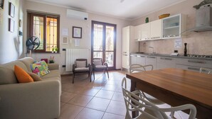 Un salon chaleureux avec un canapé, des fauteuils, une table à manger en bois et un coin cuisine équipé de placards et d’un ventilateur.