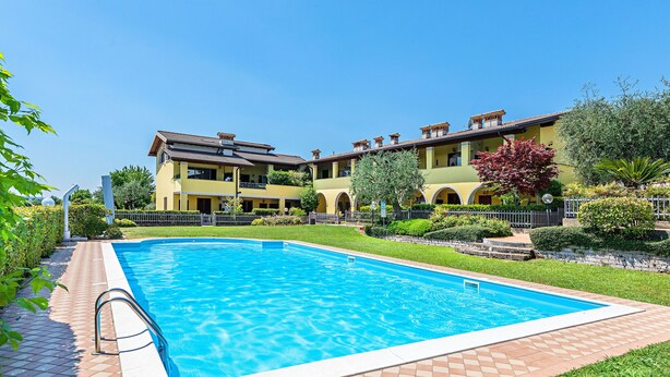 Une piscine dotée d’une terrasse carrelée, entourée d’un jardin bien entretenu et bordée d’un immeuble à plusieurs étages avec des balcons.