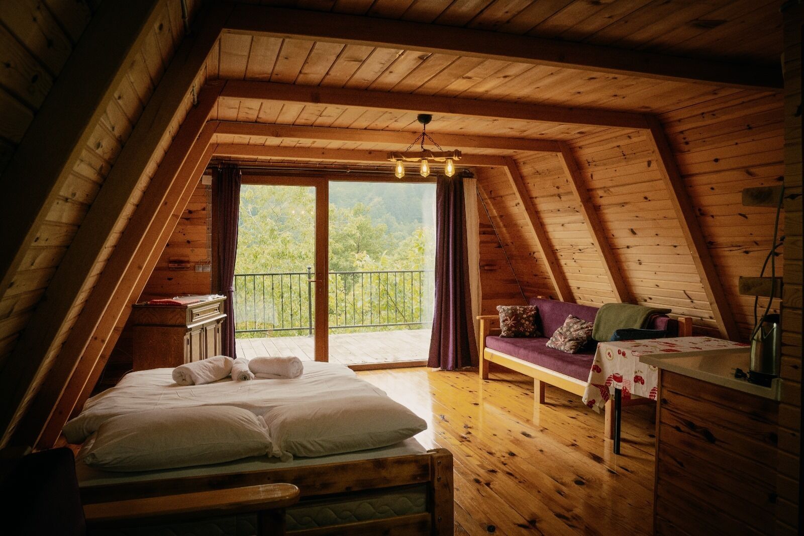 A cozy attic bedroom with a bed, a sofa, a desk, and a balcony with a view of greenery.