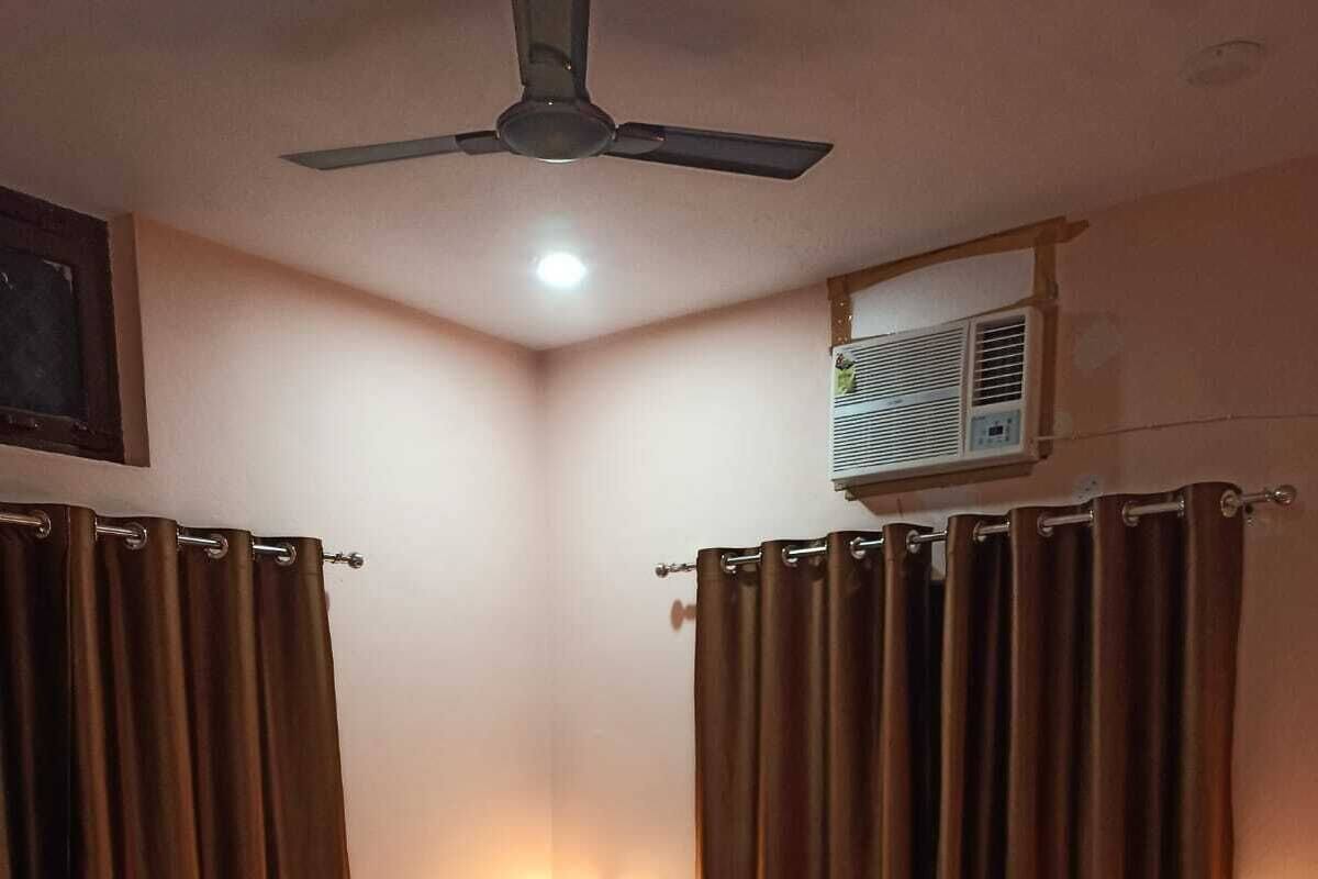 A hotel room with a bed, curtains, a ceiling fan, an air conditioning unit, and a wall-mounted light.