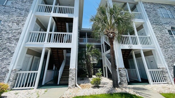A two-story building with balconies, a palm tree, and a pathway leading to the entrance. - Lovely 1BR condo in Myrtlewood (Myrtle Beach)