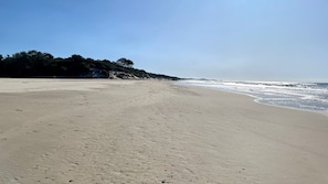 A wide sandy beach with gentle waves, a clear sky, and a forested hill in the background.