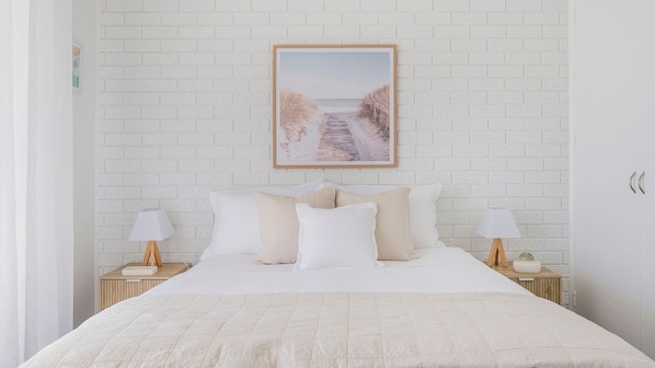 A bedroom with a white brick wall, a framed picture, a bed with white bedding, two bedside tables with lamps, and a wardrobe.