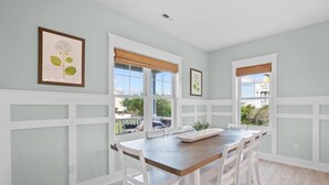 A dining area with a wooden table and white chairs, large windows with blinds, and framed artwork on the walls. - 2min Walk to Beach w Hot Tub, Centrally Located! (Oak Island)