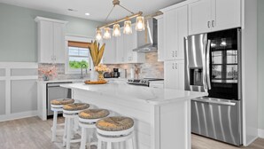 A modern kitchen with white cabinetry, a stainless steel refrigerator, and a central island with bar stools. - 2min Walk to Beach w Hot Tub, Centrally Located! (Oak Island)